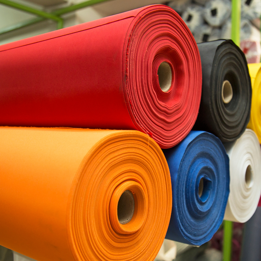 Close-up photo of several large rolls of acoustically transparent fabric stacked horizontally in different colors, including red, orange, blue, black, and white. The rolls are wound around cardboard cores, highlighting fabric thickness, smooth texture, and available color options used for acoustic panels, speakers, and sound treatment projects.