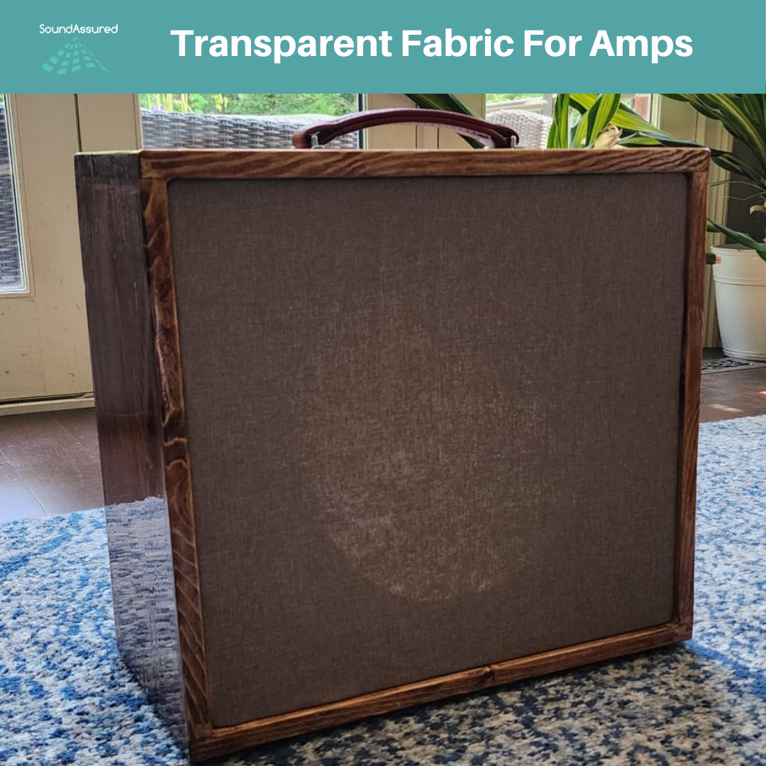A wooden guitar amplifier cabinet with a brown transparent fabric grille sits on a blue patterned rug in a well-lit room with plants in the background. A turquoise banner at the top reads “Transparent Fabric For Amps” with the SoundAssured logo on the left.